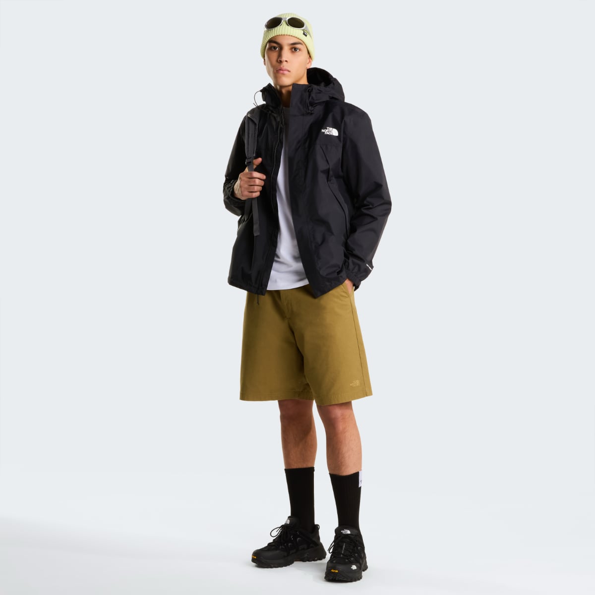 Male model in black The North Face Antora waterproof jacket with hood, backpack strap, khaki shorts and hiking boots.