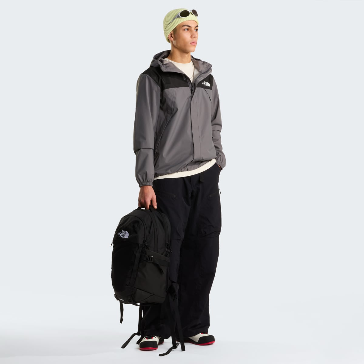 Full body shot of a male model wearing The North Face Antora waterproof hooded jacket in gray and black, with lime green beanie and black backpack.