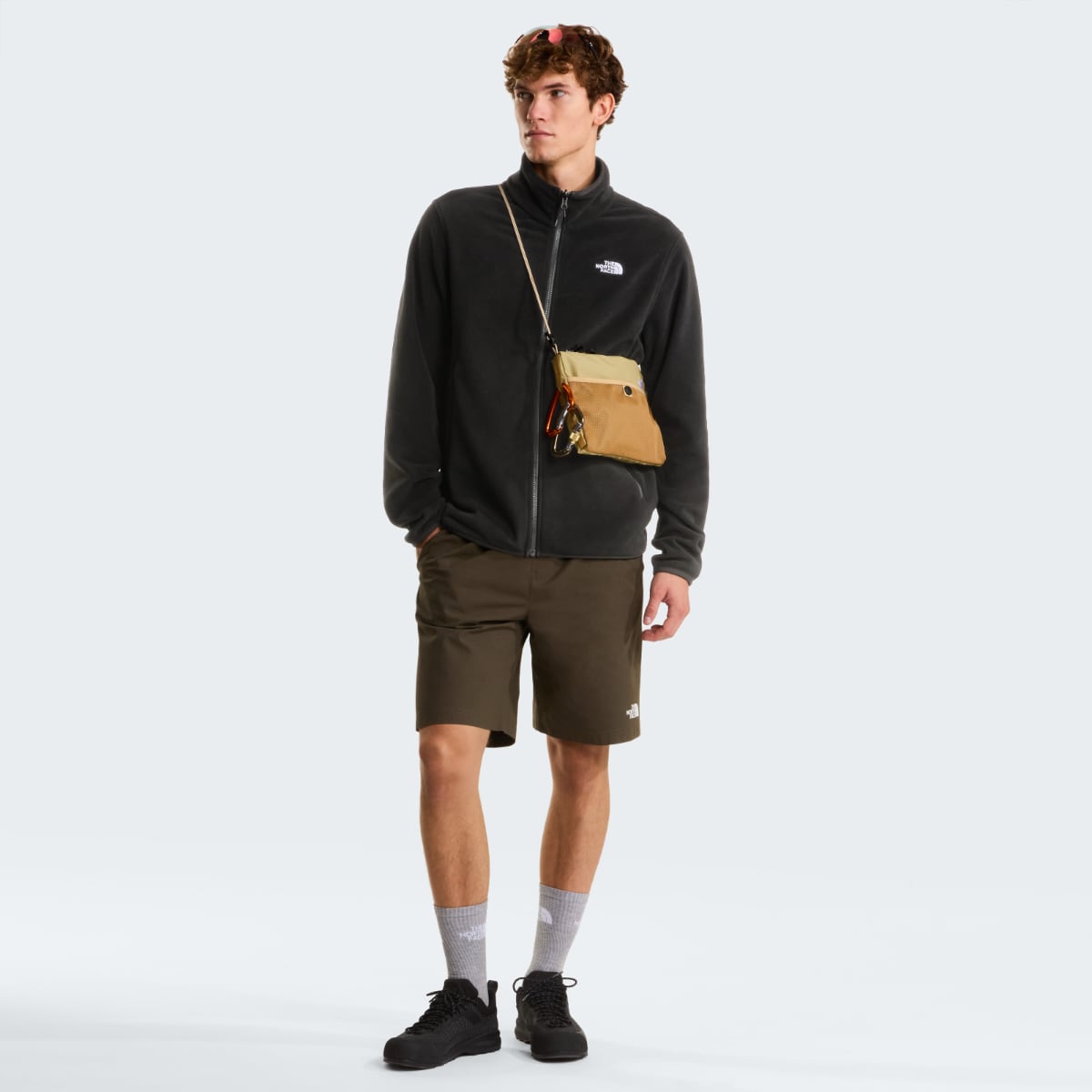 Male model in black The North Face Glacier Fleece jacket with full zip and small white logo, paired with brown shorts and a tan crossbody pouch