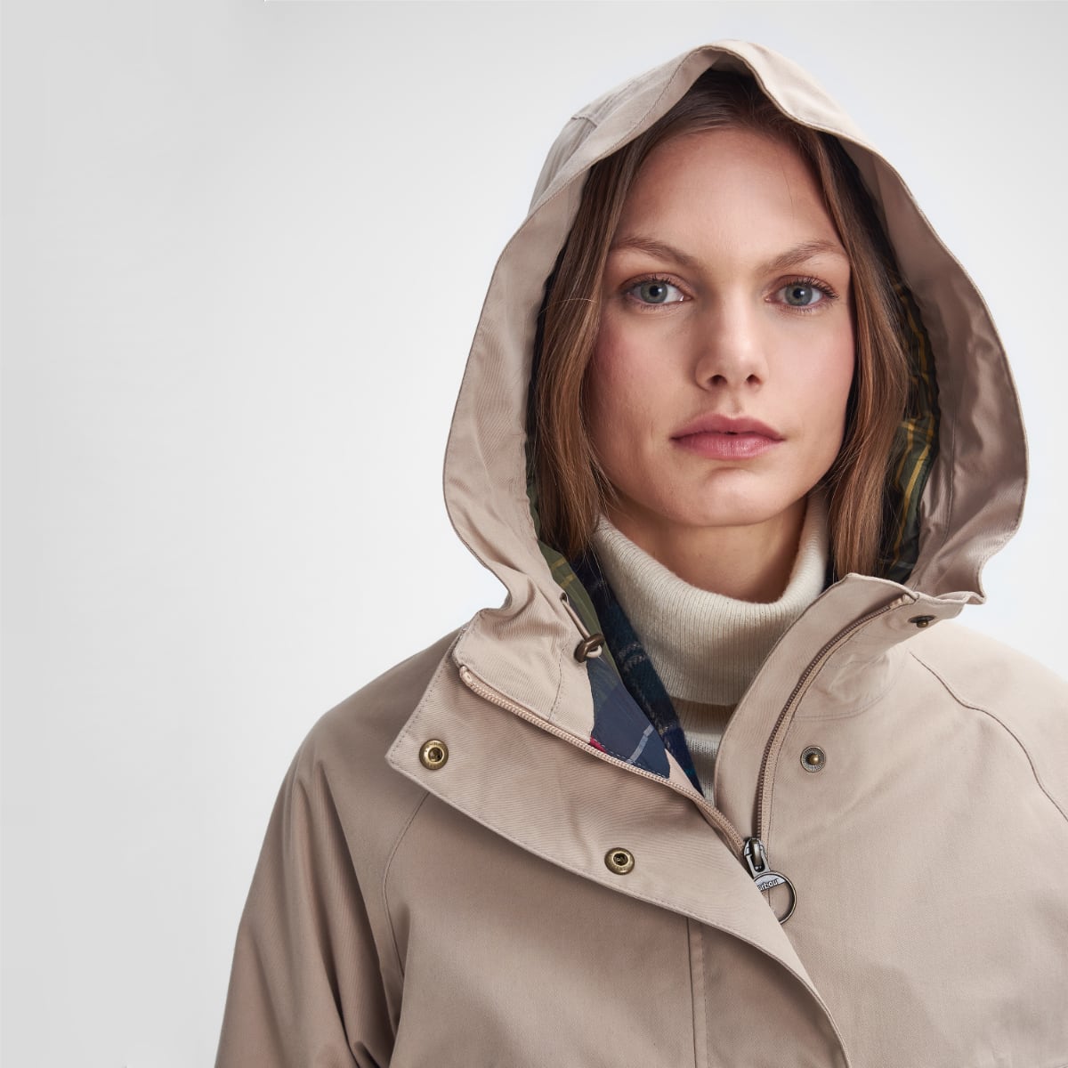 Model wearing a beige Barbour waterproof jacket with hood up and plaid lining visible at the neck.