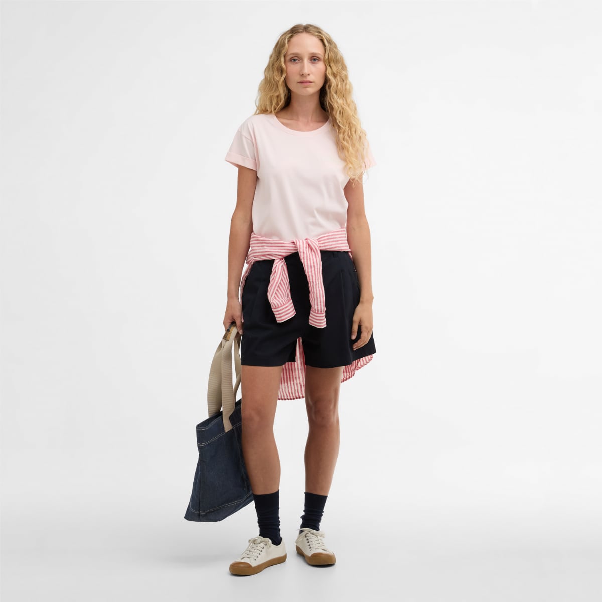 Model wearing pink Barbour Bede T Shirt with a striped pink sweater tied at the waist and navy shorts carrying a denim tote