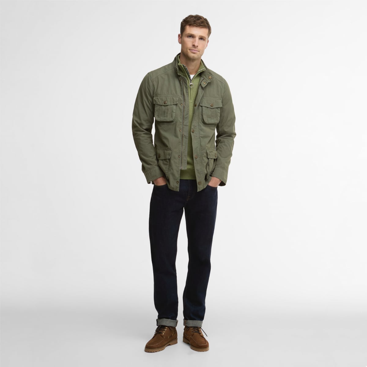 Full body view of a man in dusty olive Barbour Corbridge Utility jacket over a green sweater with dark jeans and brown hiking boots