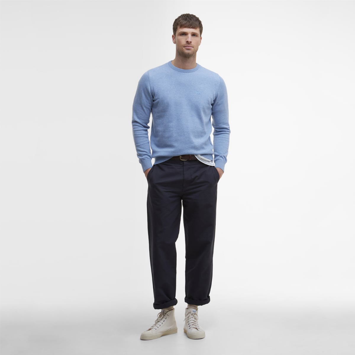 Full length image of a male model in a light blue Barbour crew neck jumper with dark navy trousers and white sneakers