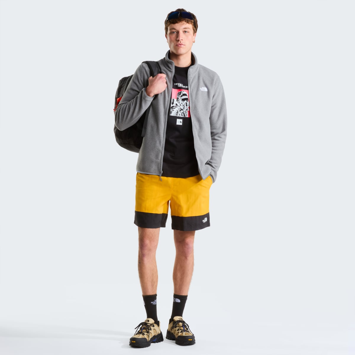 Full body shot of a man wearing grey The North Face Glacier Fleece jacket over a black graphic tee, with a backpack and yellow and black shorts