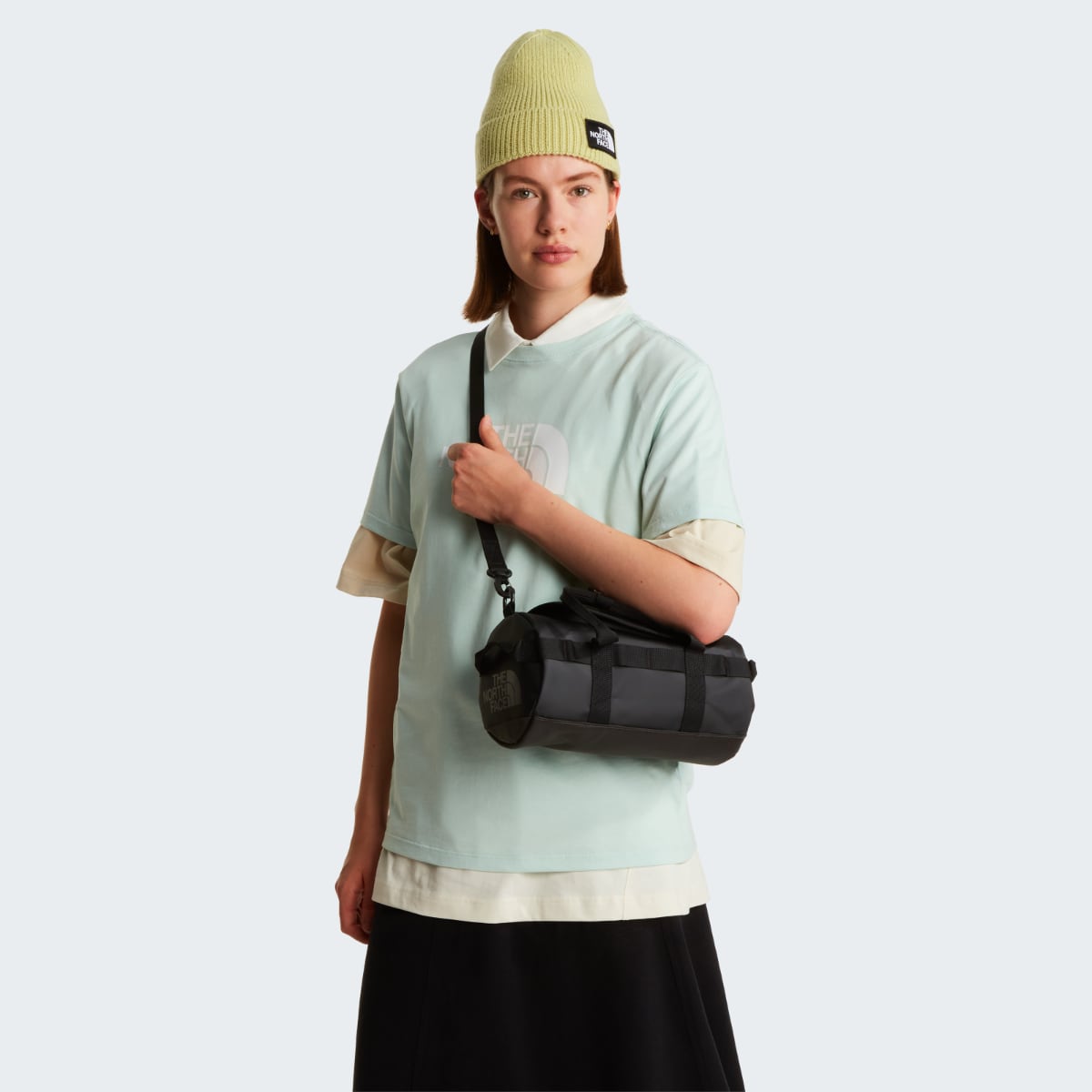 Model wearing a light mint shirt carries a compact black and grey Base Camp Duffel shoulder bag with a crossbody strap.