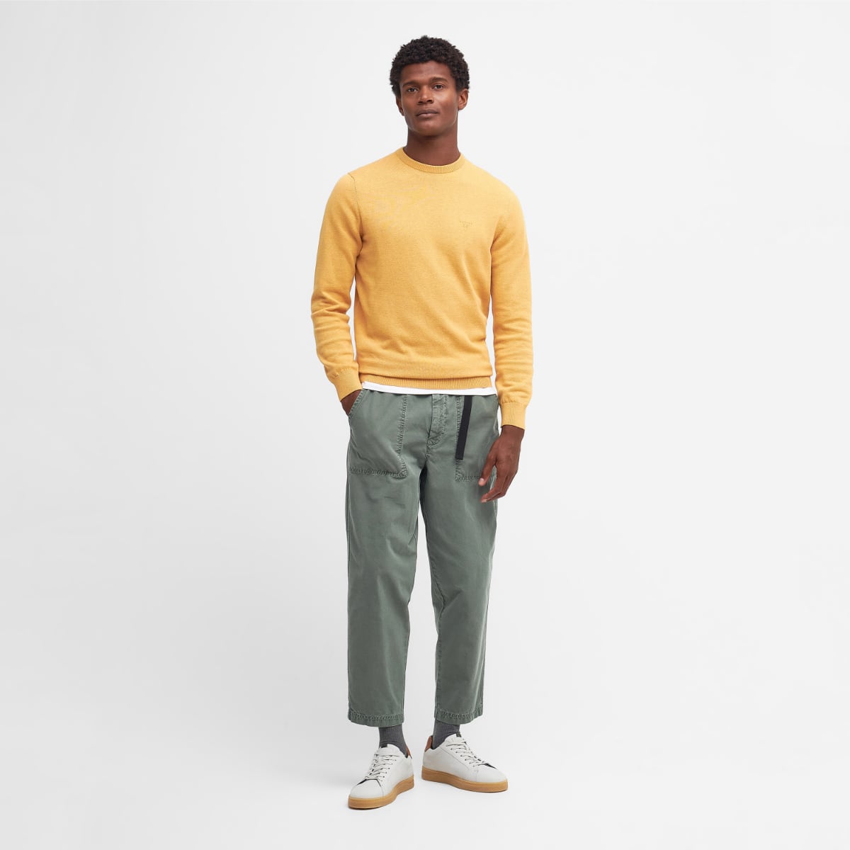 Full-body front view of a male model wearing a honey gold Barbour Pima Cotton crew neck jumper with a white tee underneath and green pants.