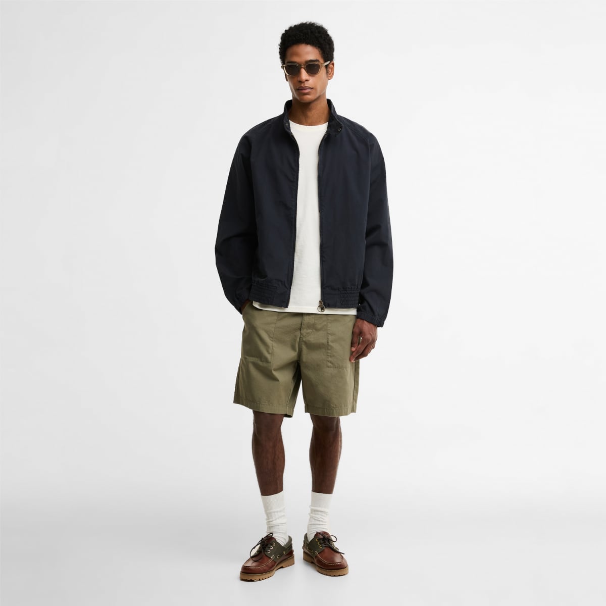 Model wearing dark navy Barbour Royston Harrington over a white tee with olive shorts and sunglasses