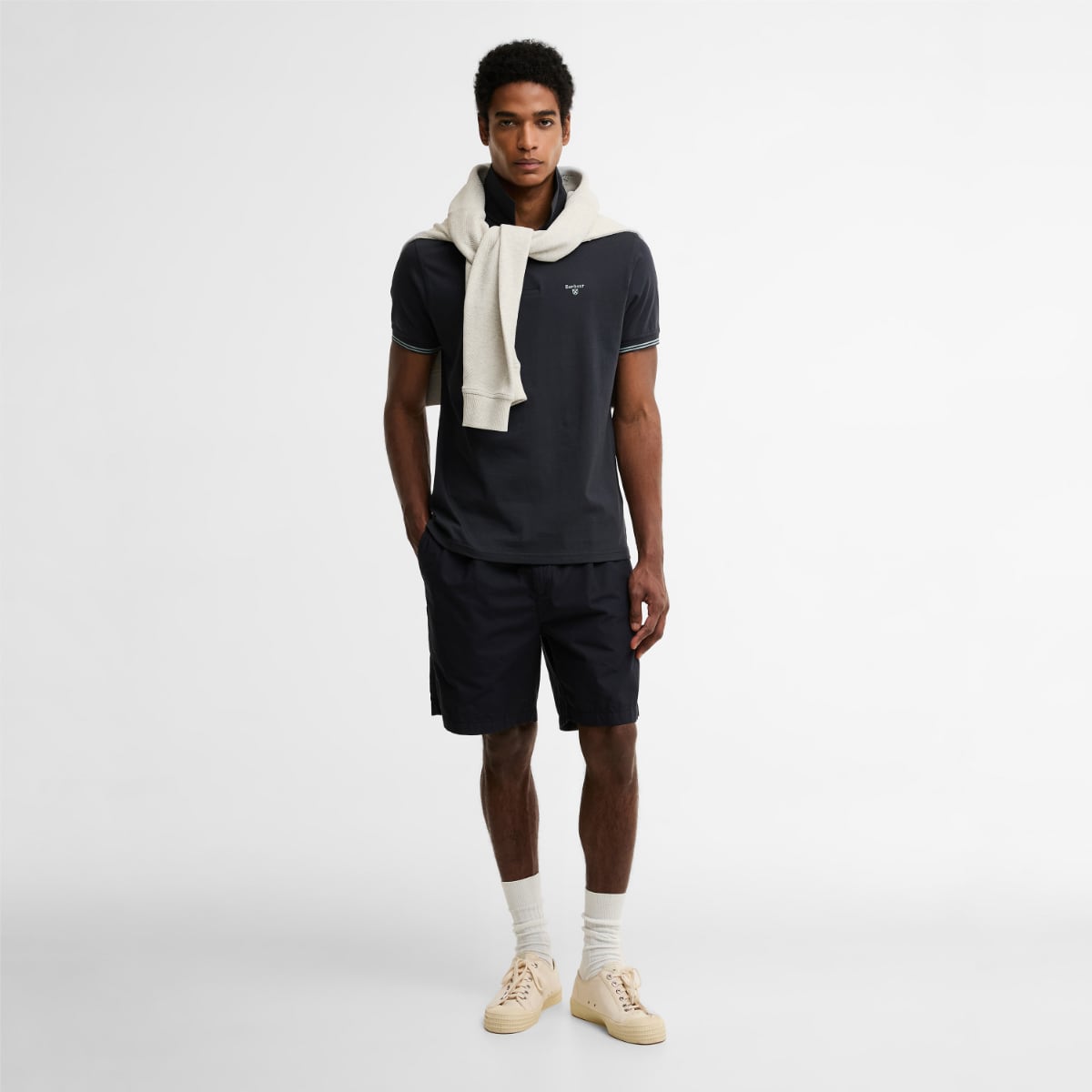 Full body shot of a male model in a dark navy Barbour Austwick polo with a cream sweater draped over shoulders