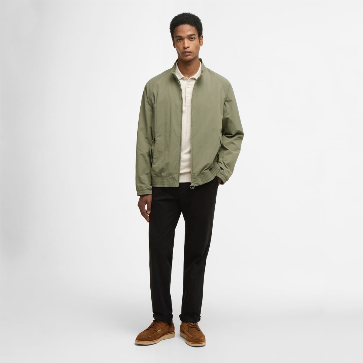Full-body shot of a male model wearing Barbour Royston Showerproof jacket in dusty olive over a beige top and black trousers.