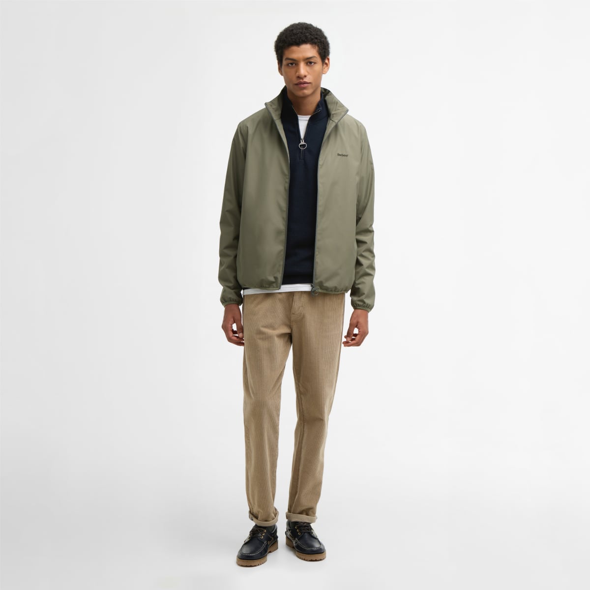 Male model wearing dusty olive Barbour Korbel waterproof jacket with zip front and standing collar, paired with navy top and tan corduroy pants.